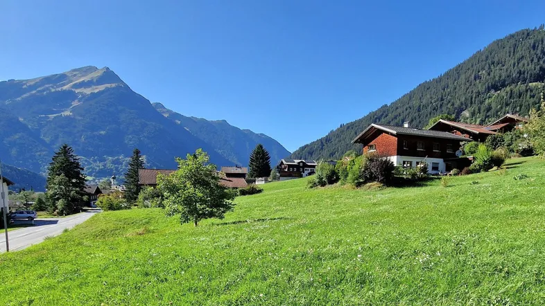 Häuser in alpiner Landschaft mit grünen Wiesen und Bergen unter blauem Himmel, sonniger Tag.