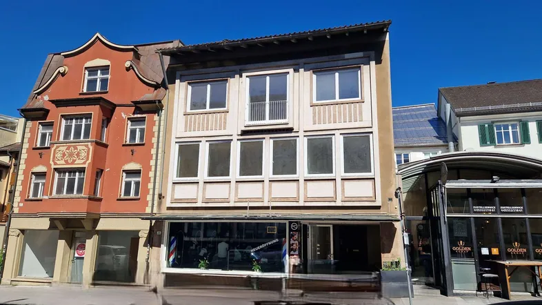 Fassade eines Mehrfamilienhauses mit Geschäften im Erdgeschoss und Wohnungen darüber an einem sonnigen Tag.