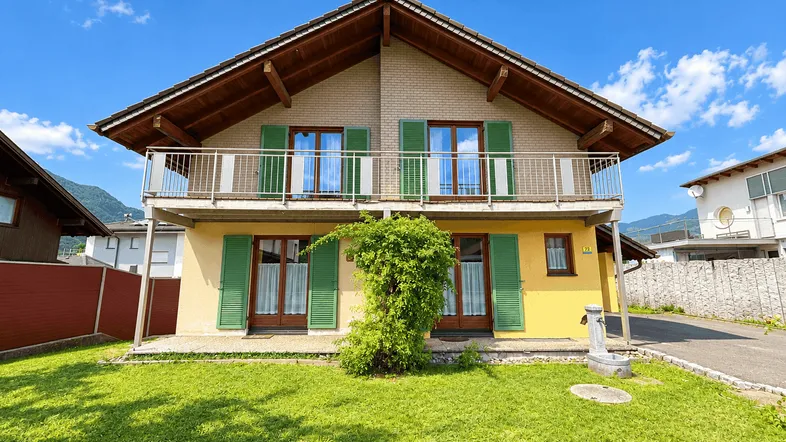 Charmantes Haus mit gelber Fassade, grünem Garten, Balkon und Bergblick unter blauem Himmel.