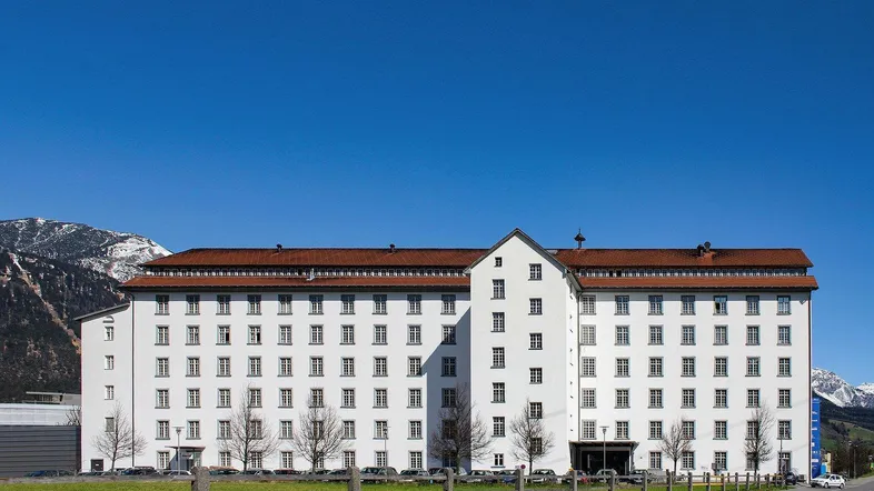 Großes weißes Gebäude mit vielen Fenstern und braunem Dach, umgeben von Bergen und einer Straße.