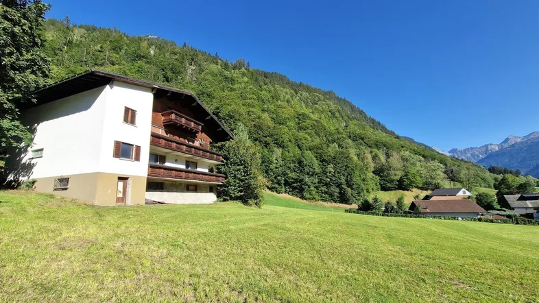 Traditionelles Haus mit Holzbalkonen und weißer Fassade, eingebettet in eine grüne Berglandschaft.