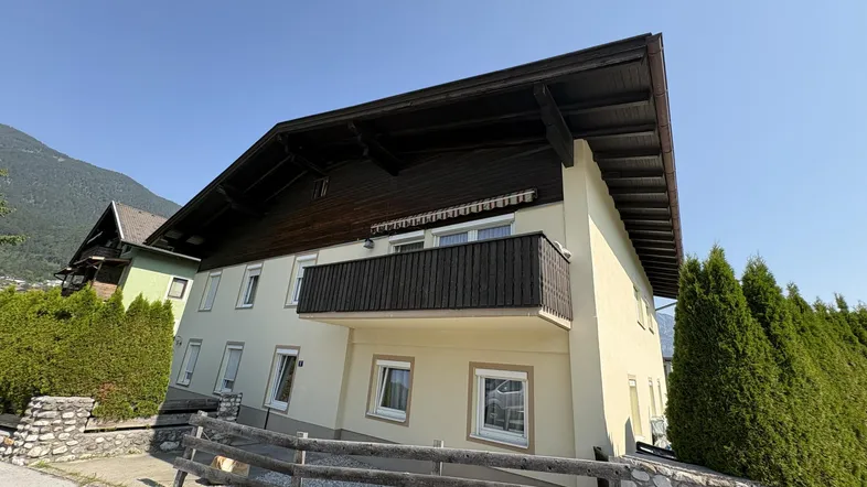 Hausfassade mit Balkon und gepflegten Hecken, umgeben von einer bergigen Landschaft.