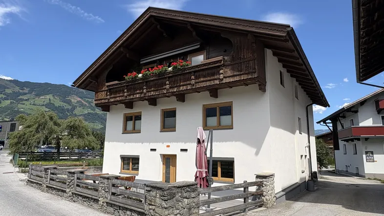 Traditionelles Haus mit weißer Fassade, Holzbalkon und Blick auf die umliegenden Berge.