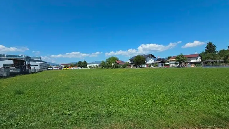 Weitläufiges grünes Feld unter blauem Himmel mit Wohnhäusern und Industriegebäuden im Hintergrund.