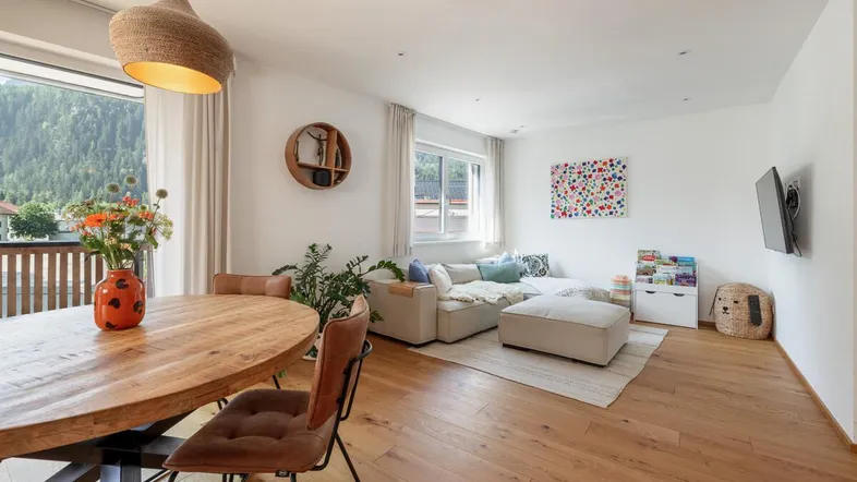 Heller Wohn- und Essbereich mit Holzboden, gemütlichem Sofa und Zugang zum Balkon mit Bergblick.