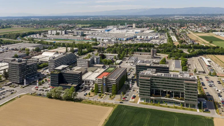 Luftaufnahme eines Gewerbegebiets mit mehreren Bürogebäuden, Parkplätzen und grünen Feldern im Vordergrund.