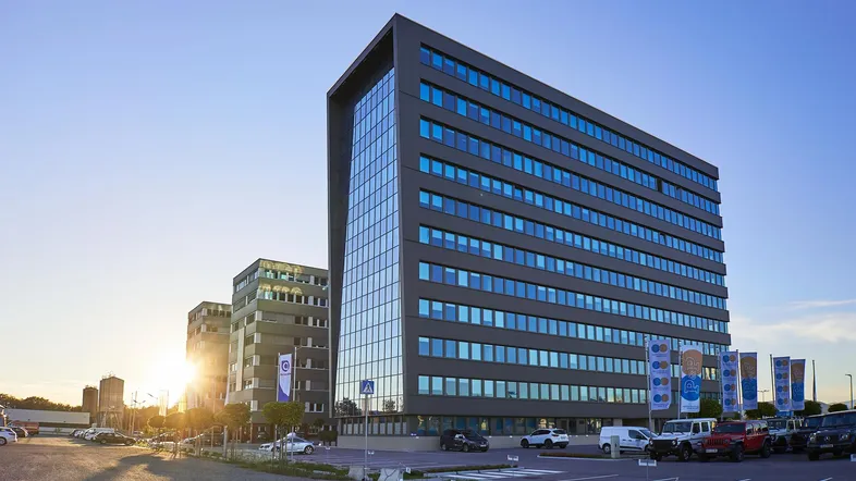 Modernes Bürogebäude mit Glasfassade und Parkplätzen bei Sonnenuntergang, umgeben von weiteren Geschäftsgebäuden.