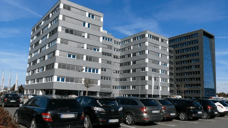 Modernes Bürogebäude mit mehreren Etagen und einer Reihe von geparkten Autos davor unter blauem Himmel.