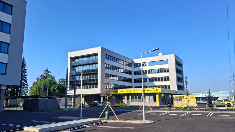 Modernes Gebäude mit großen Fenstern, Balkonen und einem Parkplatz im Vordergrund unter blauem Himmel.