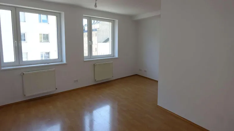Helles, leeres Zimmer mit Parkettboden und zwei Fenstern, die viel Tageslicht hereinlassen.