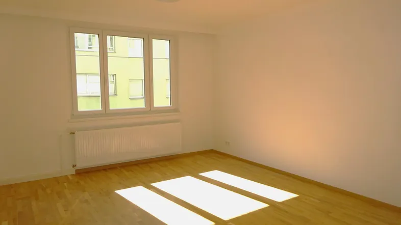 Helles, leeres Zimmer mit Parkettboden und großem Fenster, das viel Tageslicht hereinlässt.