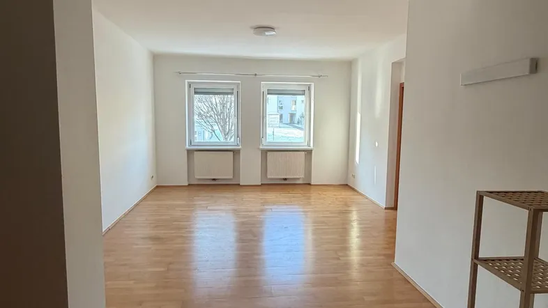 Helles, leeres Wohnzimmer mit Parkettboden und zwei Fenstern, die viel Tageslicht hereinlassen.