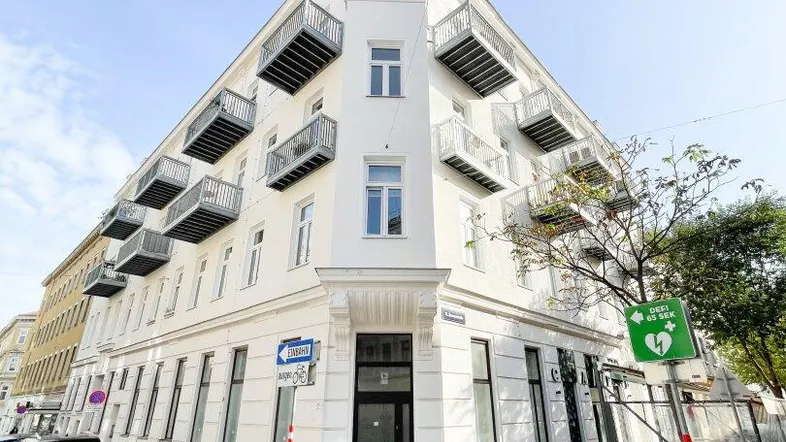 Weißes Eckgebäude mit mehreren Balkonen und Blick auf die Straße unter klarem Himmel.