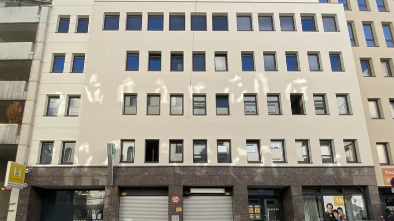 Mehrstöckiges Gebäude mit beiger Fassade, vielen Fenstern und Garagentoren im Erdgeschoss in einer städtischen Umgebung.