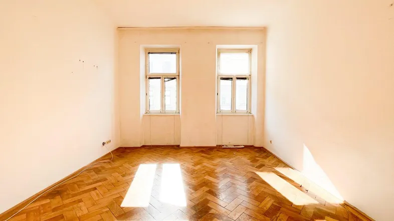 Helles Zimmer mit Fischgrätparkett und zwei Fenstern, das viel natürliches Licht hereinlässt.