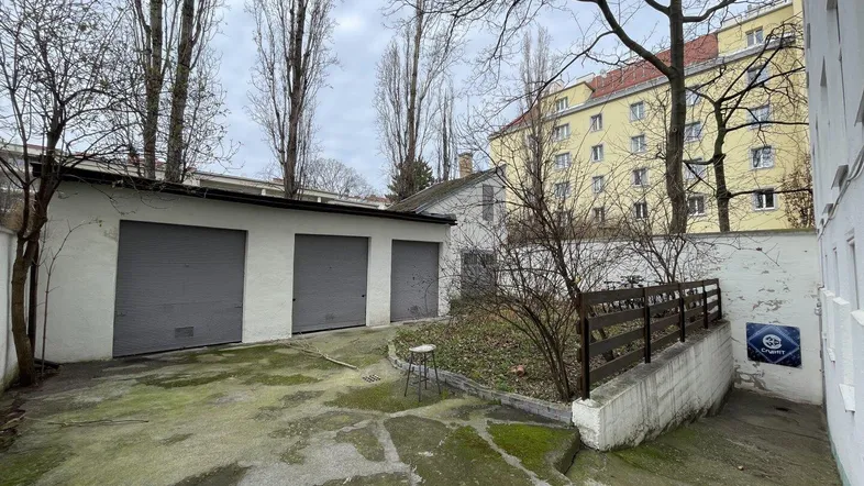 Hinterhof mit Garagen, einem kleinen Gartenbereich und hohen Bäumen im Hintergrund.
