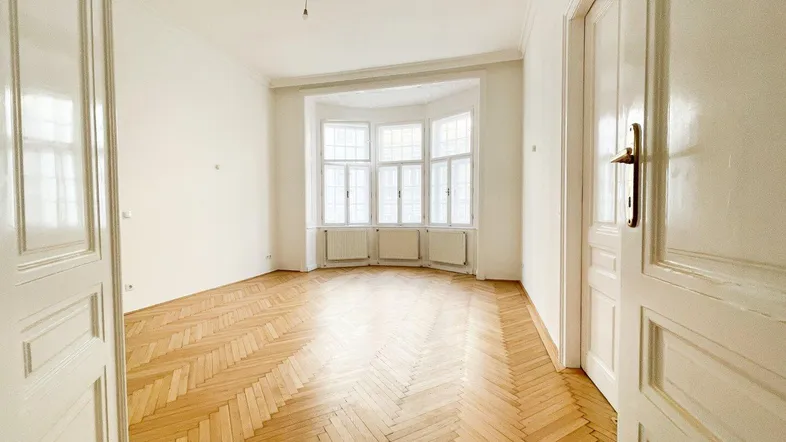 Helles, leeres Zimmer mit Fischgrätparkett und einem großen Erkerfenster, das viel Tageslicht hereinlässt.