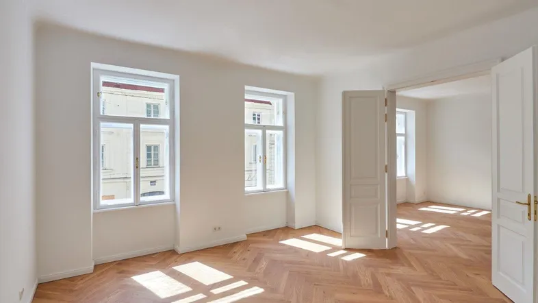 Helles Zimmer mit Fischgrätparkett, hohen Decken und viel Tageslicht durch große Fenster.