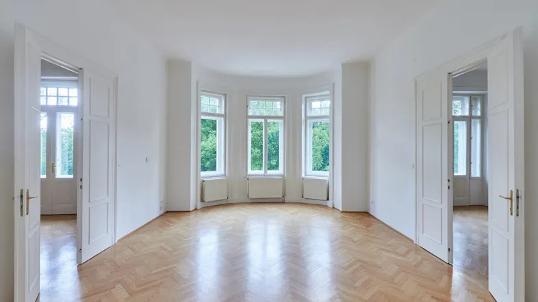 Helles, leeres Zimmer mit Fischgrätparkett und großen Fenstern mit Blick ins Grüne.