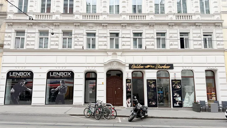 Vorderansicht eines historischen Gebäudes mit Stuckfassade und Geschäften im Erdgeschoss an einer belebten Straße.