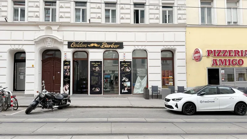 Ansicht der Straßenfassade eines Gebäudes mit Geschäften im Erdgeschoss und parkenden Fahrzeugen.