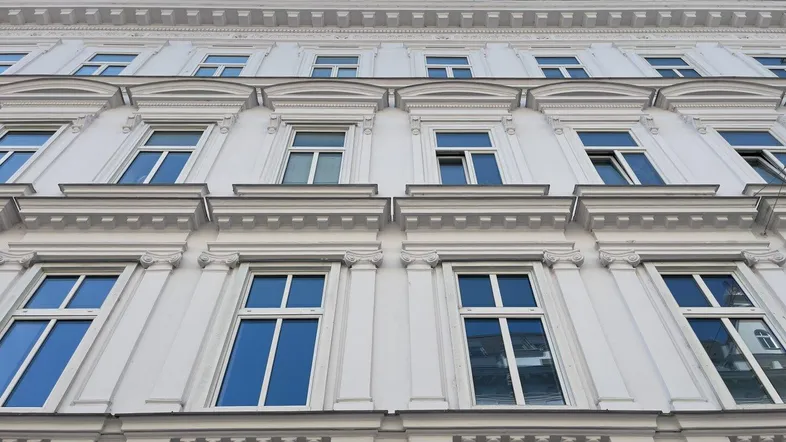 Weiße Fassade eines eleganten Altbaus mit klassischen Verzierungen, großen Fenstern und einem Mansarddach unter blauem Himmel.
