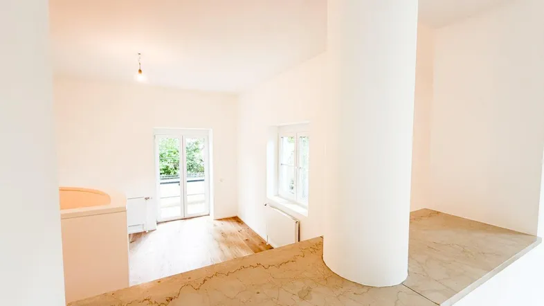 Heller Wohnbereich mit Holzfußboden, runder Säule und Zugang zu einem Balkon oder einer Terrasse.