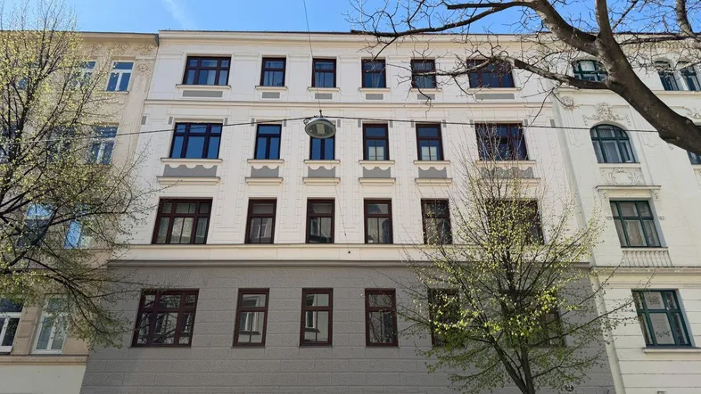 Historische Fassade eines Mehrfamilienhauses mit vielen Fenstern und Bäumen im Vordergrund.