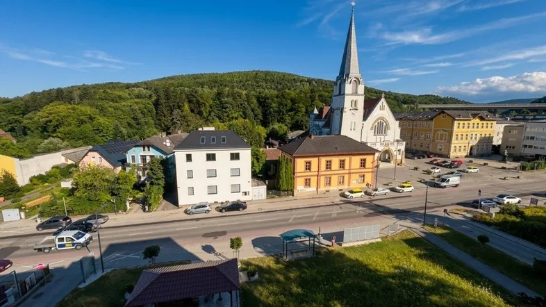 Luftaufnahme einer Stadt mit Kirche, Häusern und grünen Hügeln unter blauem Himmel.