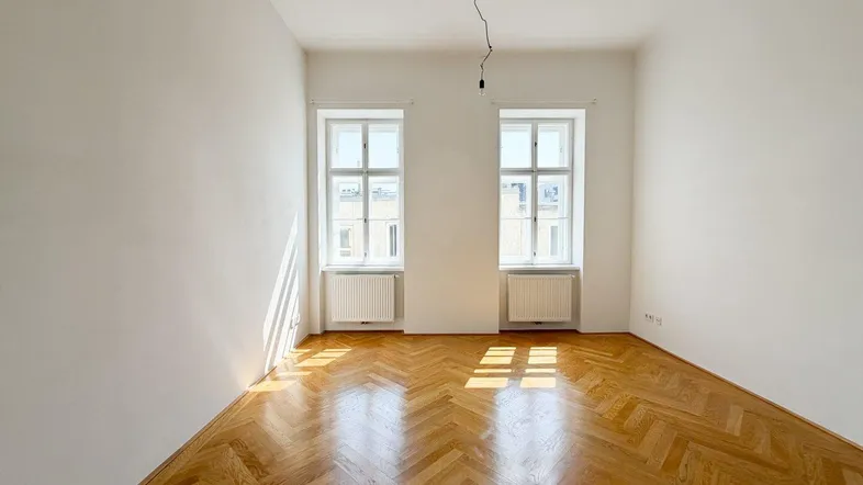 Helles Zimmer mit Fischgrätparkett und zwei Fenstern, das viel Tageslicht hereinlässt.