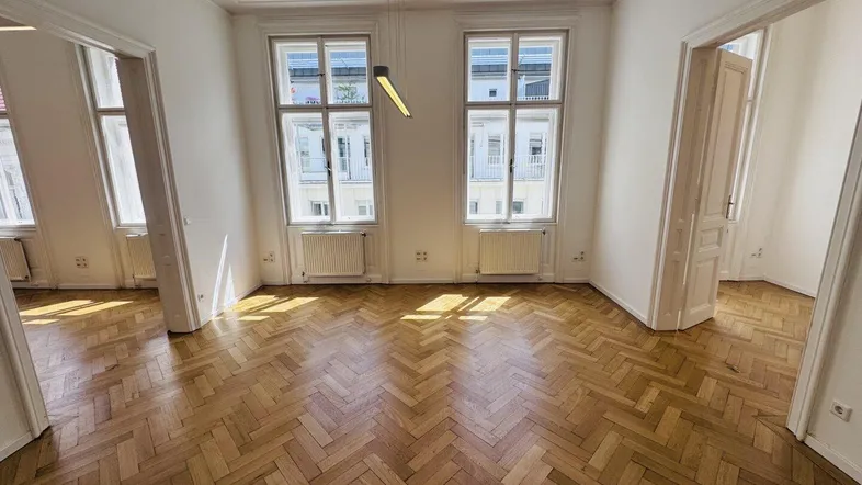 Großes Wohnzimmer mit klassischem Fischgrätparkett, hohen Decken und einer modernen Deckenleuchte.