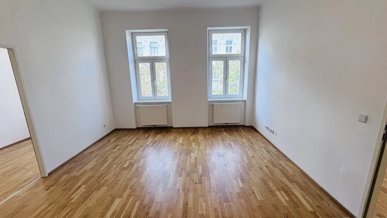 Helles, leeres Zimmer mit Parkettboden und zwei Fenstern, bereit zur individuellen Gestaltung.