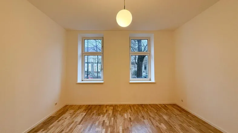 Heller Wohnraum mit Holzboden und zwei Fenstern, die viel Tageslicht hereinlassen.