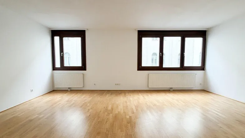 Helles, leeres Zimmer mit Parkettboden, weißen Wänden und zwei großen Fenstern.