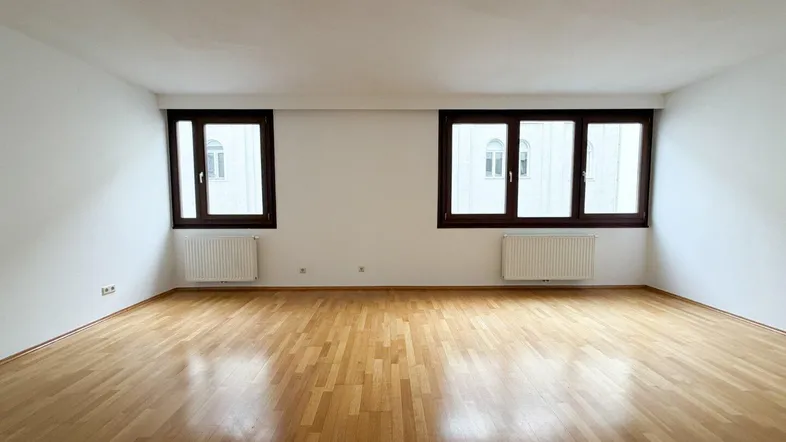 Helles, leeres Wohnzimmer mit Parkettboden und zwei Fenstern, bereit zur individuellen Gestaltung.
