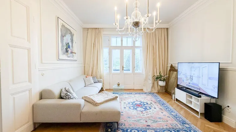 Helles Wohnzimmer mit elegantem Kronleuchter, Fischgrätparkett und Zugang zu einem Balkon.