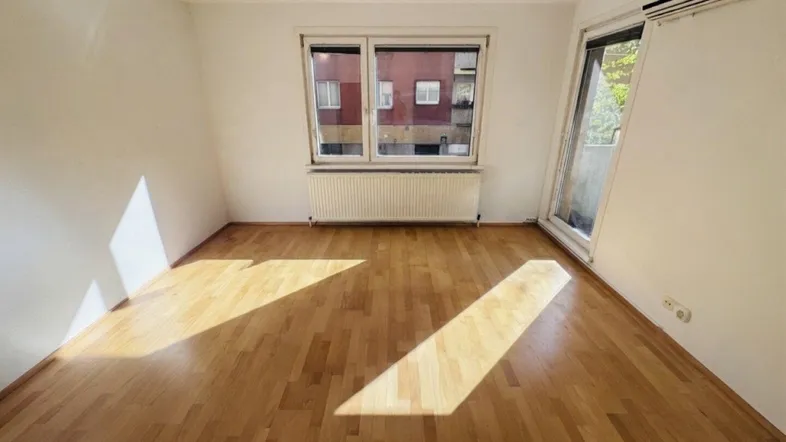 Helles, leeres Zimmer mit Parkettboden und Fenster, das viel Sonnenlicht hereinlässt.