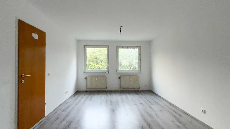 Helles, leeres Zimmer mit Laminatboden und zwei Fenstern, die einen Blick ins Grüne bieten.