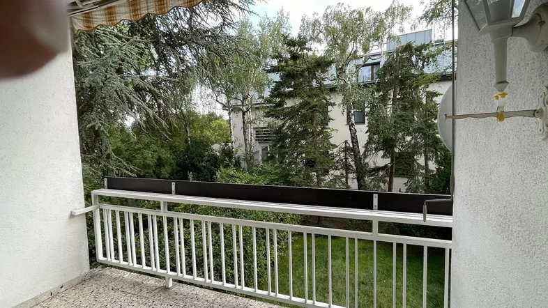 Balkon mit Markise und Blick ins Grüne, umgeben von Bäumen und anderen Gebäuden.