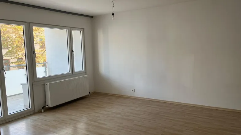 Helles, leeres Zimmer mit Laminatboden, weißen Wänden und Zugang zu einem Balkon.