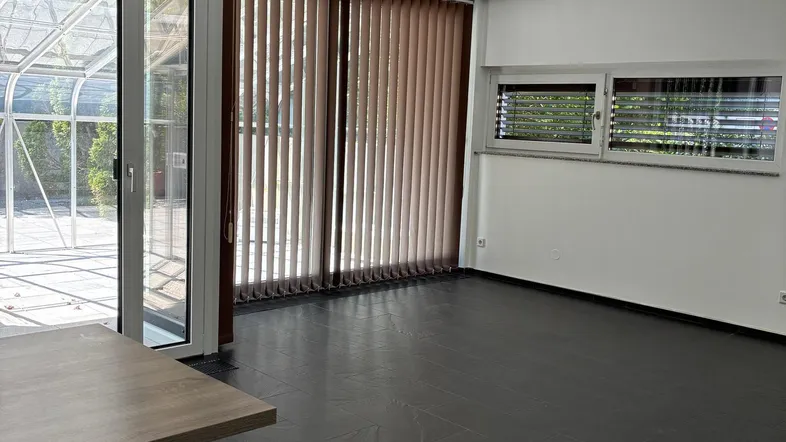 Helles Wohnzimmer mit dunklen Fliesen, großen Fenstern und Zugang zu einem Wintergarten.