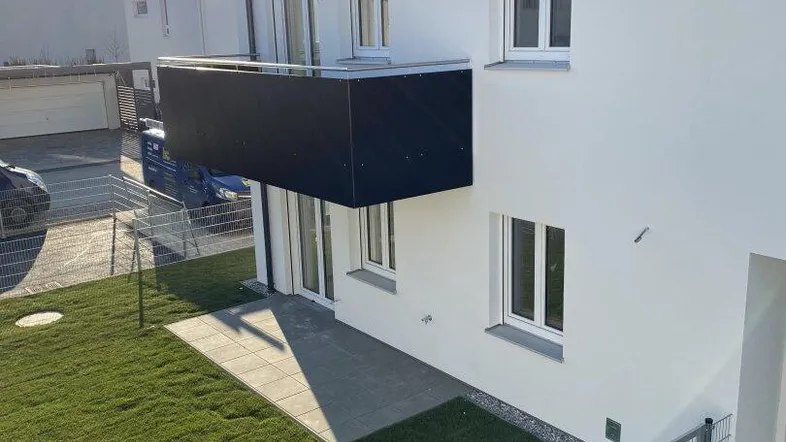 Modernes Gebäude mit weißer Fassade, kleinem Gartenbereich und schwarzem Balkon unter strahlender Sonne.