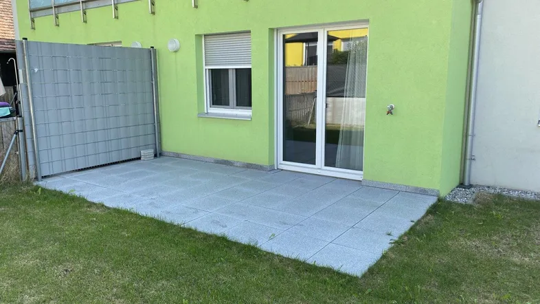 Gepflasterte Terrasse mit Rasenfläche und grün gestrichener Hausfassade, ideal für Entspannung.