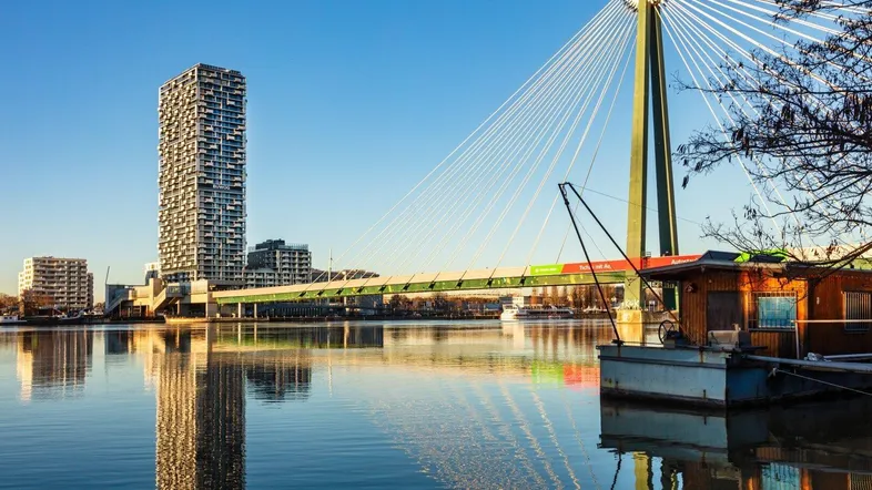 Panoramablick auf ein modernes Hochhaus am Wasser mit einer markanten Brücke im Vordergrund unter blauem Himmel.