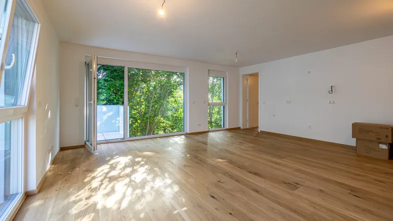 Helles, leeres Wohnzimmer mit Holzboden und Zugang zu einem kleinen Balkon mit Blick ins Grüne.