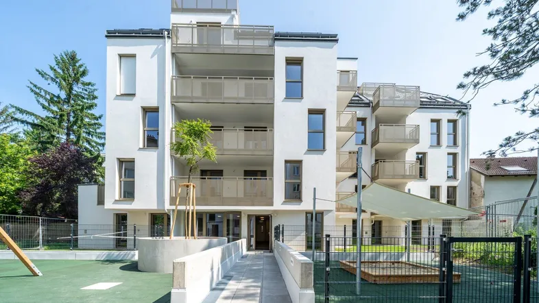 Modernes Mehrfamilienhaus mit weißer Fassade, Balkonen und einem Spielplatz im Vordergrund.