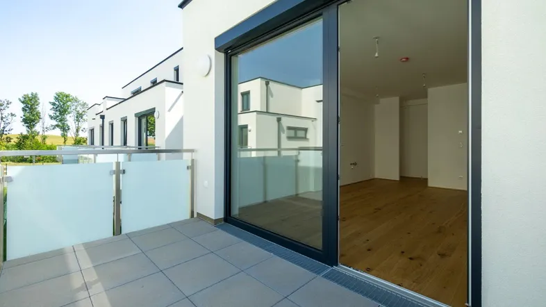 Heller Balkon mit Fliesenboden und Glasgeländer, mit Blick in einen leeren Raum.