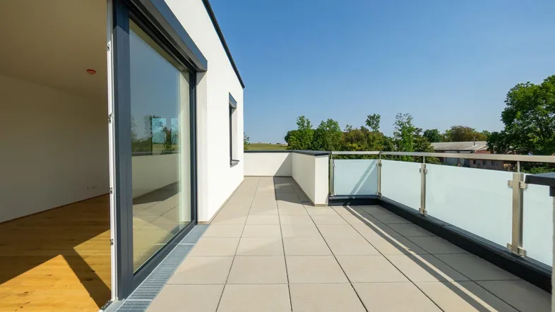 Große Dachterrasse mit modernen Fliesen, Glasgeländer und Zugang zu einem hellen Innenraum.