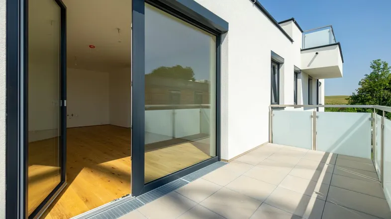 Heller Balkon mit Fliesenboden und Glasgeländer, bietet Zugang zu einem lichtdurchfluteten Innenraum.