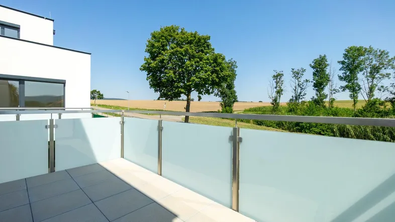 Moderne Terrasse mit Fliesenboden und Glasgeländer, bietet einen weiten Blick ins Grüne.